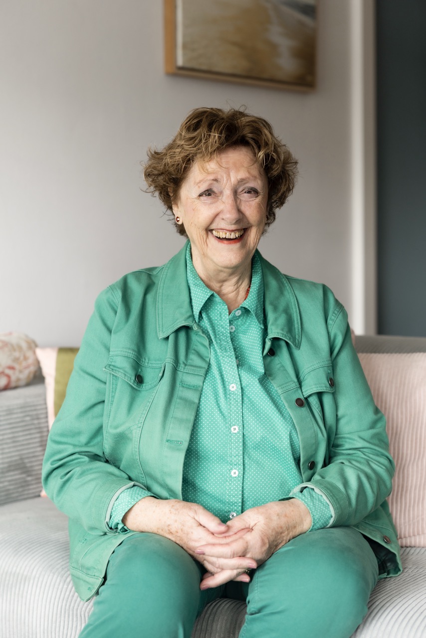 Een vrouw zit op de bank van haar huis en lacht naar de camera.