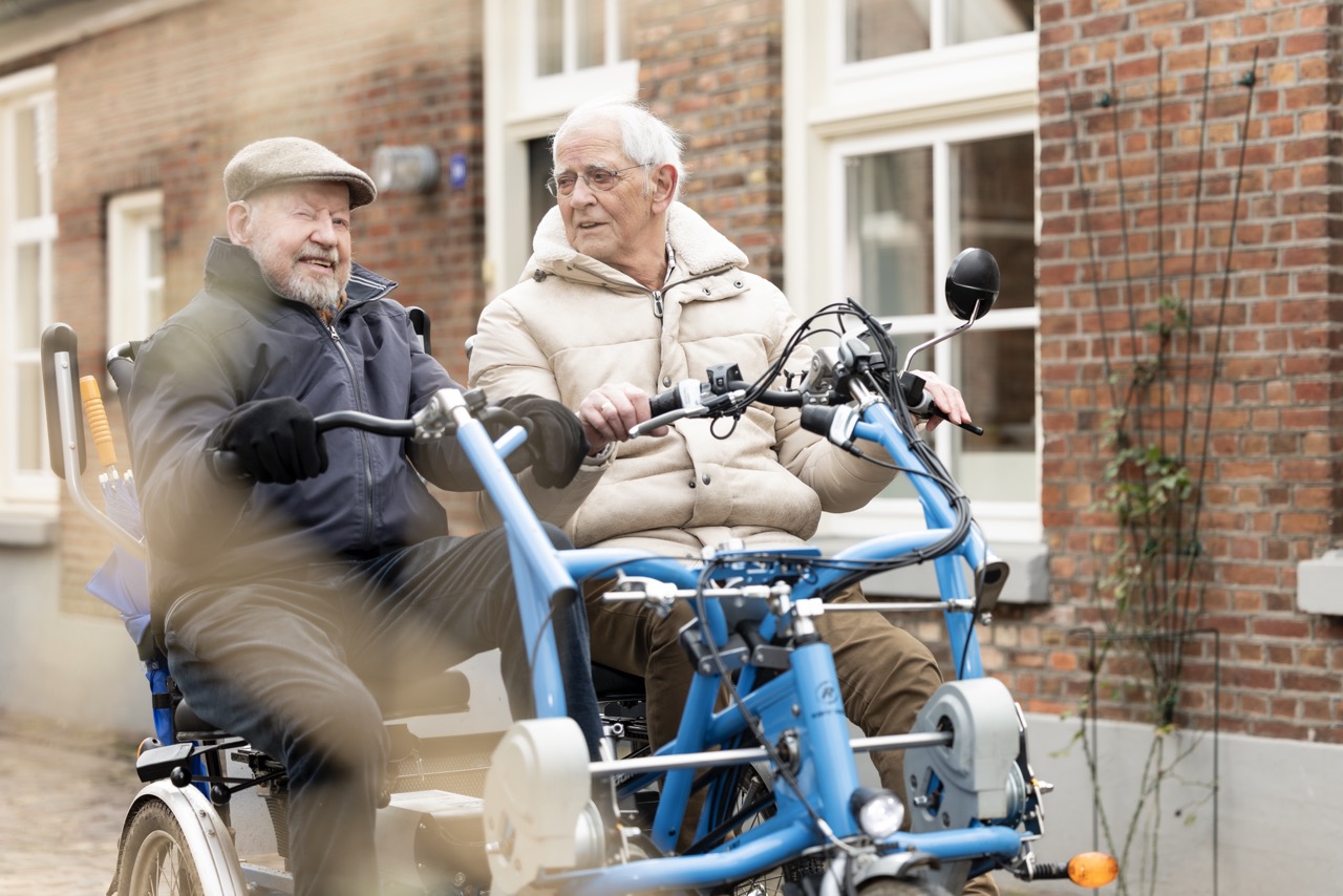 Twee mannen zitten samen op een duofiets. Één van hen kijkt naar de ander.