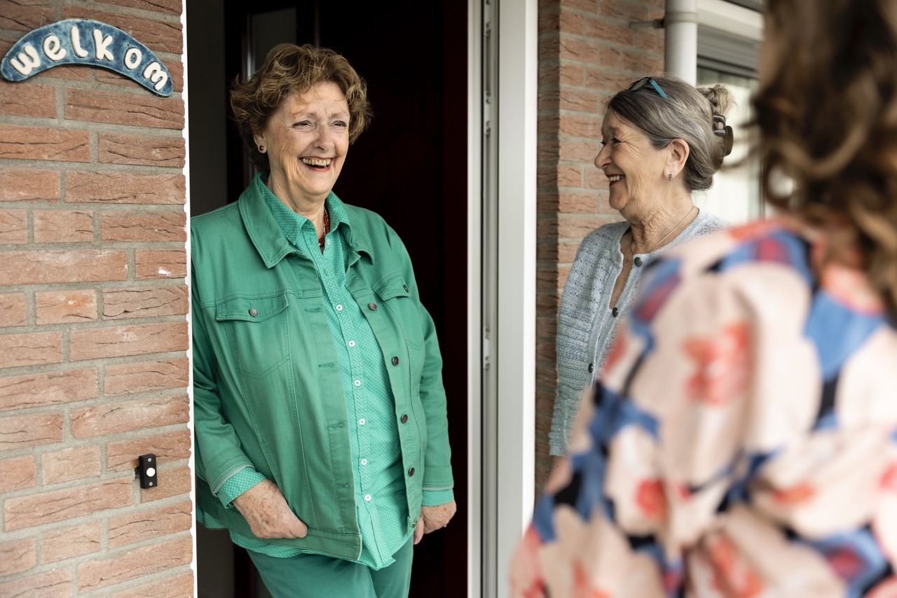 Een vrouw staat in de opening van haar deur. Een andere vrouw staat naast haar en lacht naar haar.
